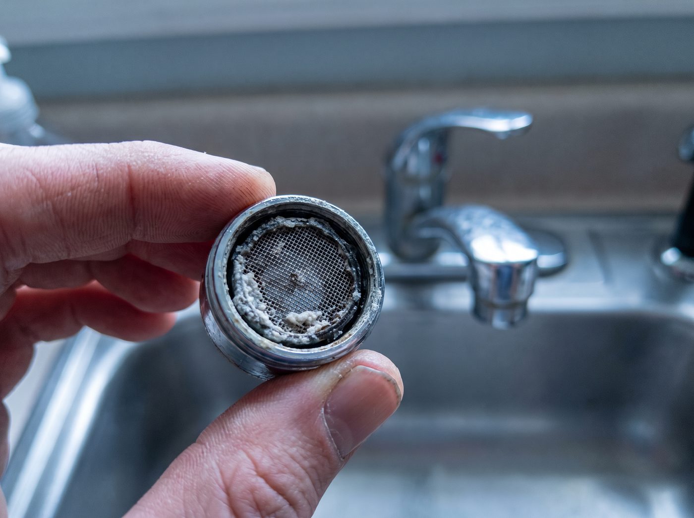 A faucet aerator held in fingertips with its mesh screen packed with mineral scale
